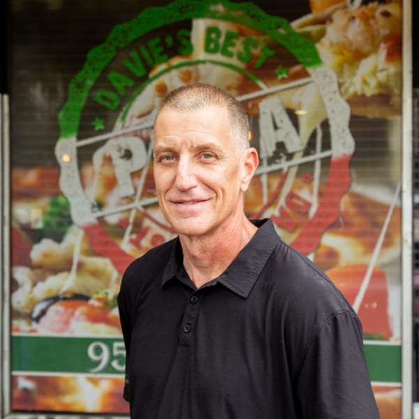 A man in a black polo shirt smiles in front of Davie's Best Pizza Bar window.