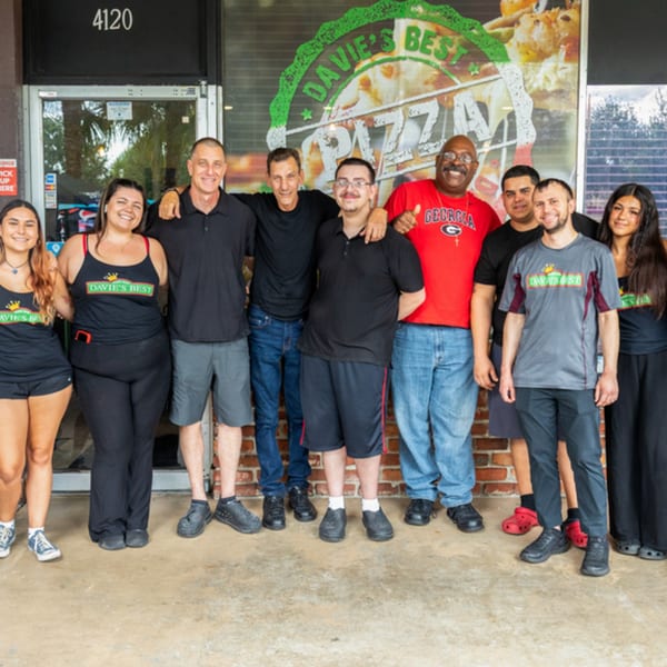 Nine smiling Davie's Best Pizza Bar employees stand together in front of the restaurant's entrance.