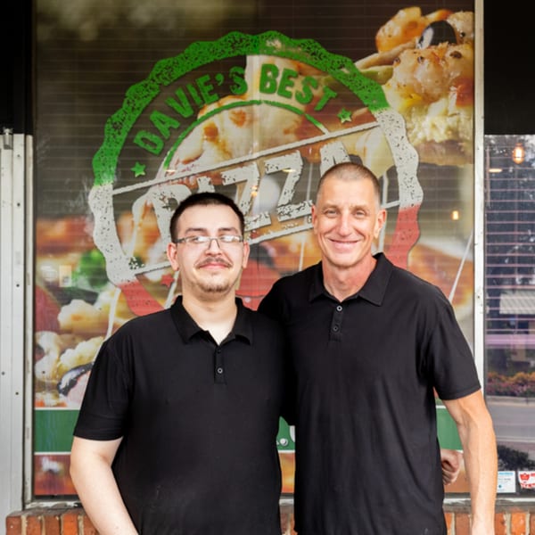 Two men in black shirts smiling in front of the "Davie's Best Pizza" storefront.